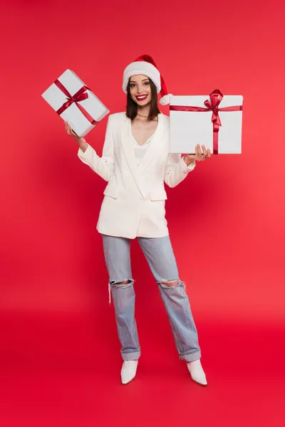 Smiling woman in santa hat holding gift boxes on red background — Stock Photo