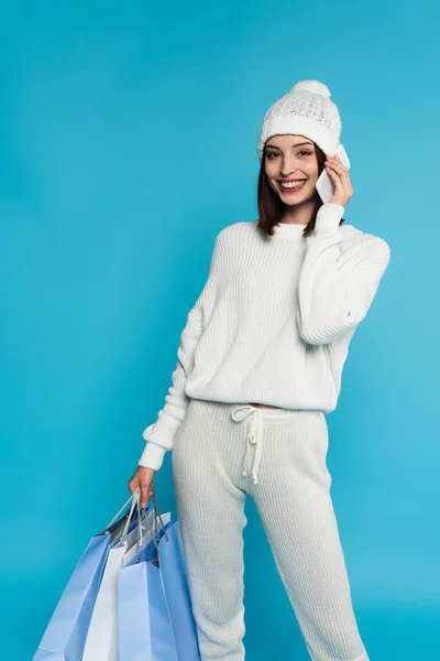 Mulher positiva no chapéu falando no smartphone e segurando sacos de compras isolados no azul — Fotografia de Stock