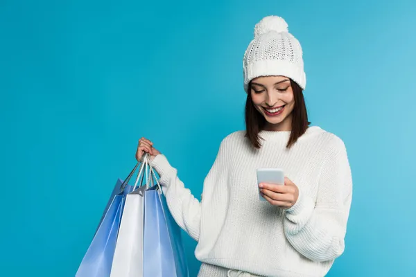 Mujer joven en ropa de punto usando teléfono inteligente y sosteniendo bolsas de compras aisladas en azul - foto de stock