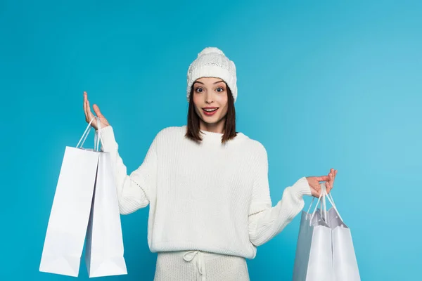 Mulher excitada em chapéu de malha e suéter segurando sacos de compras isolados em azul — Fotografia de Stock