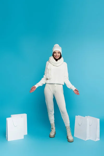 Positive Frau in Strickkleidung zeigt auf Einkaufstüten auf blauem Hintergrund — Stockfoto