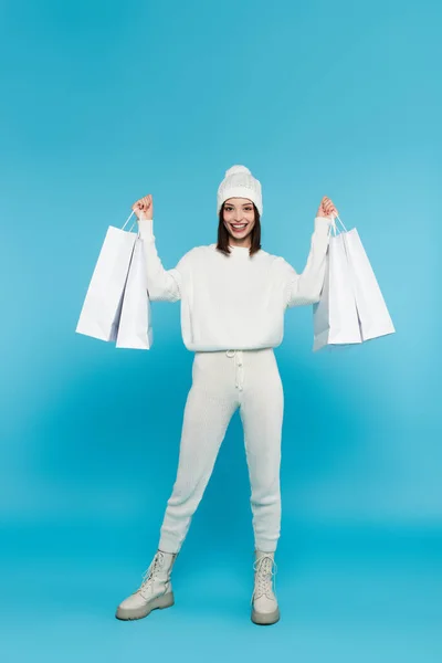 Shopaholic alegre em suéter e chapéu segurando sacos de compras no fundo azul — Fotografia de Stock