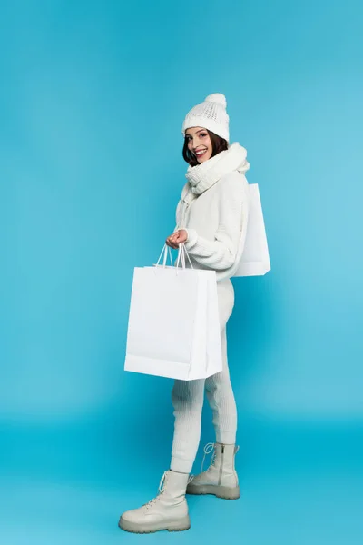 Pleine longueur de femme souriante dans un chapeau chaud et écharpe tenant des sacs à provisions sur fond bleu — Photo de stock