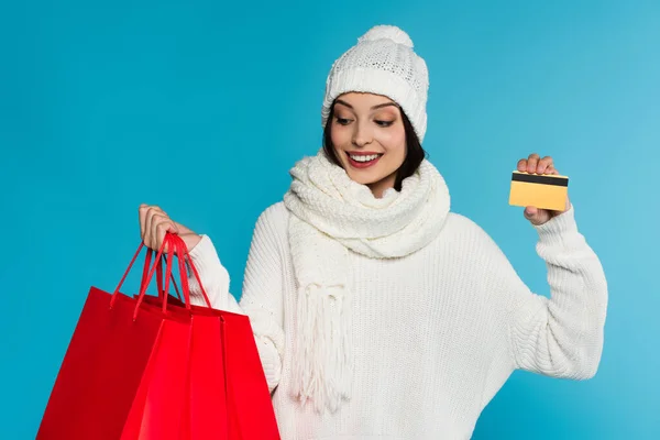 Mujer sonriente con ropa de abrigo sosteniendo bolsas rojas y tarjeta de crédito aislada en azul - foto de stock