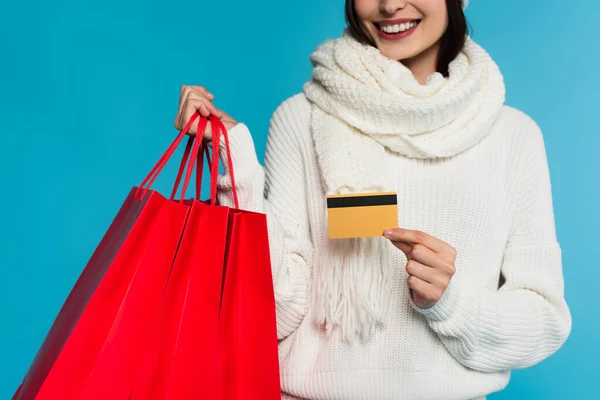 Vista recortada de una mujer sonriente en suéter con tarjeta de crédito y bolsas rojas aisladas en azul - foto de stock