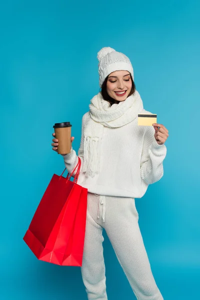 Jolie femme en tricot tenant des sacs à provisions, carte de crédit et tasse en papier isolé sur bleu — Photo de stock