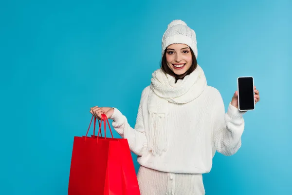 Mujer joven en ropa de abrigo sosteniendo teléfono inteligente con pantalla en blanco y bolsas de compras aisladas en azul - foto de stock