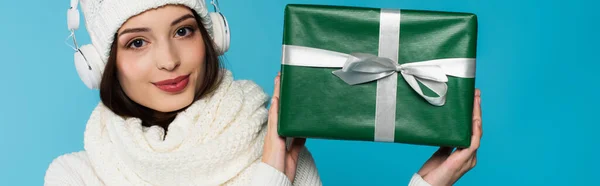 Woman in knitted scarf and headphones holding gift box isolated on blue, banner — Stock Photo