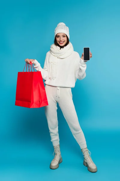 Donna sorridente in abiti a maglia che mostra smartphone con schermo bianco e shopping bag su sfondo blu — Foto stock