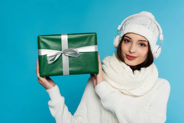 Brunette woman in headphones and warm sweater holding present with ribbon isolated on blue — Stock Photo