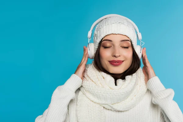 Mujer joven en auriculares y bufanda escuchando música en auriculares con los ojos cerrados aislados en azul - foto de stock