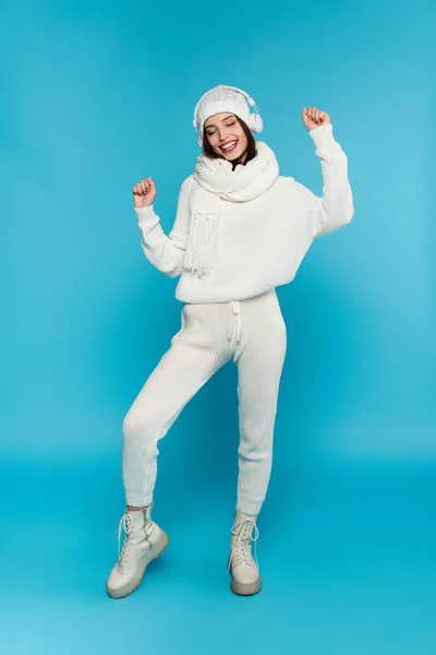 Piena lunghezza di donna sorridente in maglia cappello e maglione ballare e ascoltare musica in cuffie su sfondo blu — Foto stock