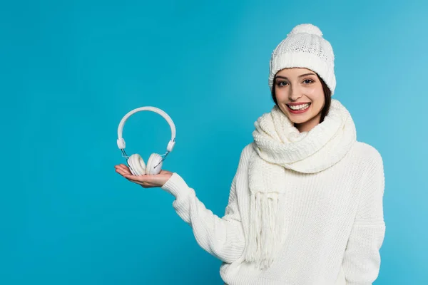 Mulher sorridente em suéter branco e chapéu segurando fones de ouvido isolados em azul — Fotografia de Stock