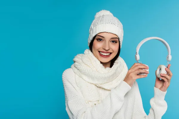 Smiling woman in knitted hat and scarf holding wireless headphones isolated on blue — Stock Photo