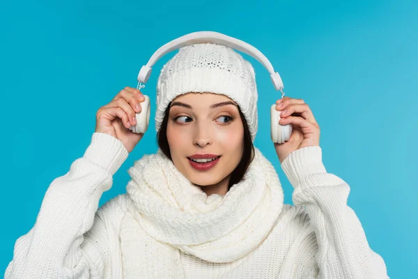 Jovem mulher de chapéu branco e suéter segurando fones de ouvido perto da cabeça isolada em azul — Fotografia de Stock