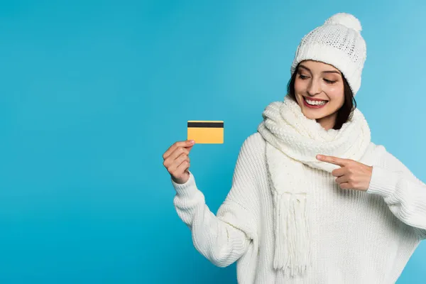 Mulher positiva em suéter quente e chapéu apontando para cartão de crédito isolado em azul — Fotografia de Stock