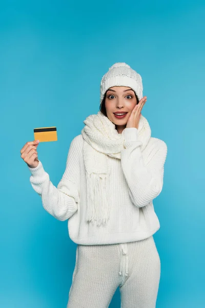 Mujer excitada en ropa de punto agarrada de la mano cerca de la mejilla y la tarjeta de crédito aislada en azul - foto de stock