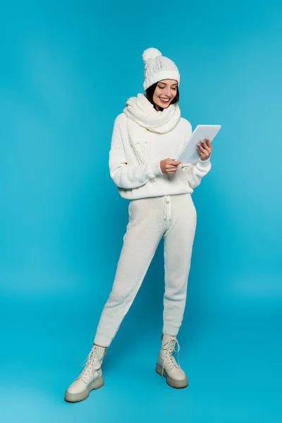 Полная длина улыбающейся женщины в теплом шарфе и шляпе, используя цифровую табличку на синем фоне — стоковое фото