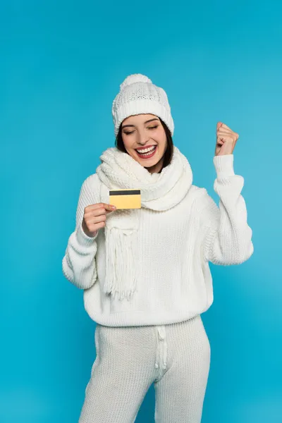 Mulher excitada em roupas brancas de malha mostrando sim gesto e segurando cartão de crédito isolado em azul — Fotografia de Stock