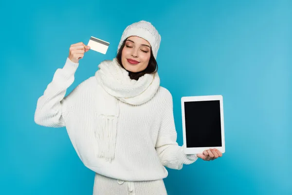 Lächelnde Frau in kuscheligem Pullover und Hut mit Kreditkarte und digitalem Tablet mit leerem Bildschirm auf blauem Hintergrund — Stockfoto