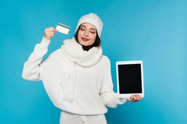 Mujer sonriente en suéter acogedor y sombrero con tarjeta de crédito y tableta digital con pantalla en blanco aislada en azul - foto de stock