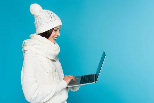 Вид сбоку улыбающегося фрилансера в вязаной шляпе и шарфе с ноутбуком, изолированным на синем фоне — стоковое фото