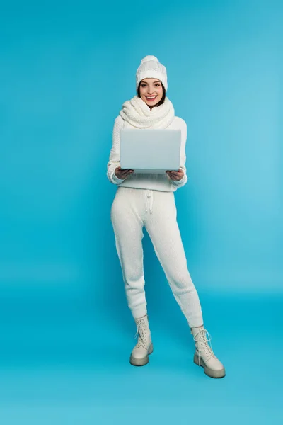 Donna sorridente in cappello a maglia che tiene il computer portatile su sfondo blu — Foto stock