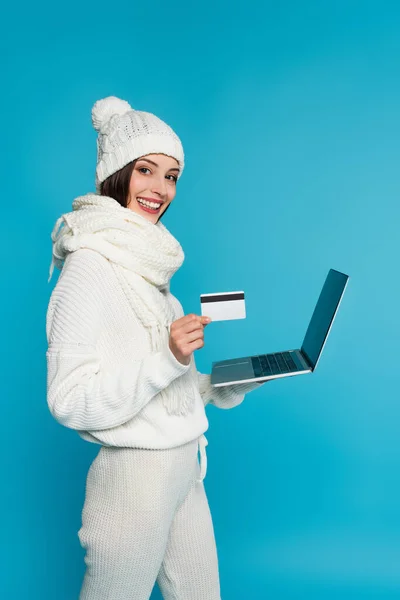 Mujer alegre en sombrero blanco que sostiene el ordenador portátil y la tarjeta de crédito aislada en azul - foto de stock