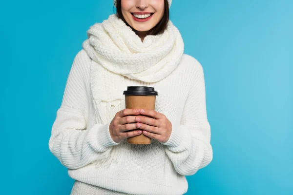 Обрізаний вид усміхненої жінки в білому в'язаному шарфі, що тримає паперову чашку ізольовано на синьому — стокове фото