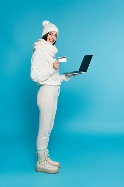 Full length of smiling woman in knitted clothes holding credit card and laptop with blank screen on blue background — Stock Photo