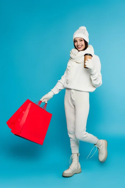 Mujer alegre en ropa de punto y guantes sosteniendo taza de papel y bolsas de compras sobre fondo azul - foto de stock