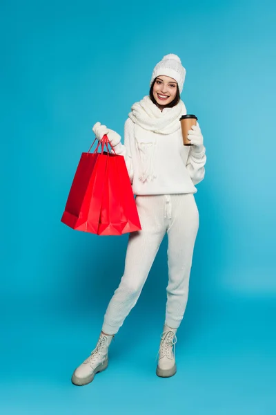 Mulher positiva em camisola de malha e luvas segurando café para ir e sacos de compras no fundo azul — Fotografia de Stock