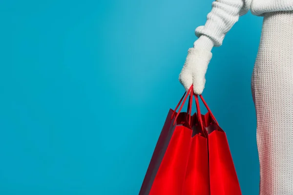 Vista recortada de la mujer en suéter y guante sosteniendo bolsas de compras rojas aisladas en azul - foto de stock