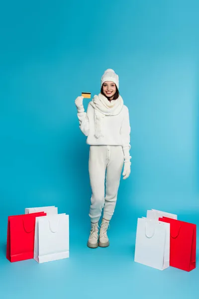 Femme souriante en vêtements chauds et gants tenant la carte de crédit près des sacs à provisions sur fond bleu — Photo de stock