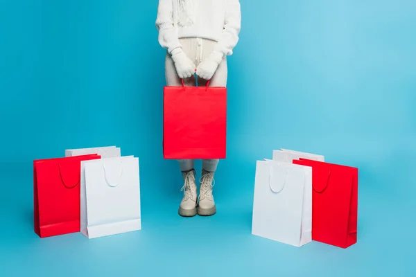 Vista cortada de mulher em suéter e luvas segurando saco de compras perto de compras em fundo azul — Fotografia de Stock