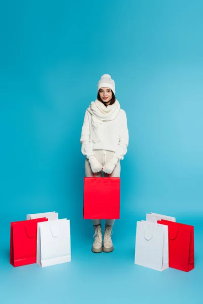 Muito shopaholic em roupas de malha de pé perto de sacos de compras no fundo azul — Fotografia de Stock