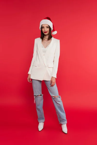 Elegante donna in cappello di Babbo Natale guardando la fotocamera su sfondo rosso — Foto stock