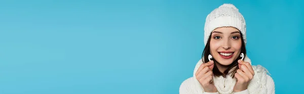 Mujer joven con ropa de abrigo sonriendo mientras sostiene auriculares inalámbricos aislados en azul, pancarta - foto de stock