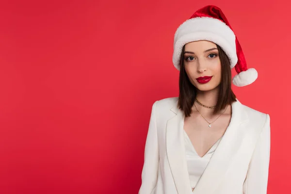 Красива жінка в капелюсі Санта і біла куртка дивиться на камеру ізольовано на червоному — стокове фото