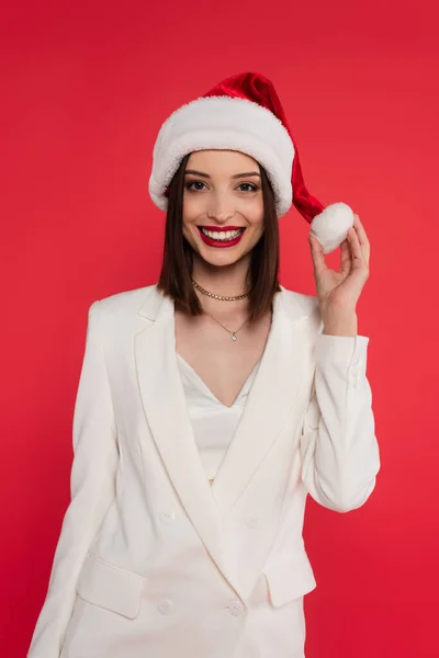 Positive woman in white jacket holding santa hat isolated on red — Stock Photo