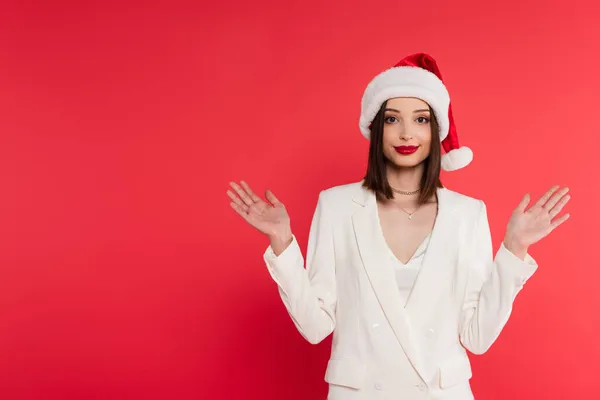 Stylische Frau mit roten Lippen und Weihnachtsmütze, die mit auf Rot isolierten Händen zeigt — Stockfoto