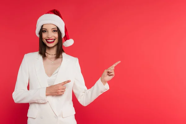 Cheerful woman in santa hat and white jacket pointing with fingers isolated on red — Stock Photo