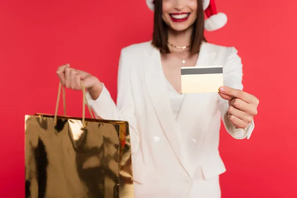 Ausgeschnittene Ansicht einer verschwommenen Frau mit Weihnachtsmann-Hut, die Einkaufstasche und Kreditkarte isoliert auf Rot hält — Stockfoto