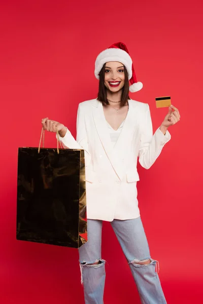Позитивна жінка в капелюсі Санта і білій куртці тримає сумку для покупок і кредитну картку ізольовано на червоному — стокове фото