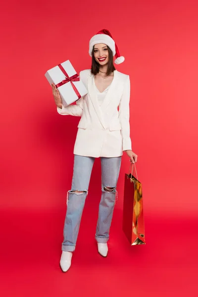 Femme gaie dans santa chapeau tenant sac à provisions et présent sur fond rouge — Photo de stock