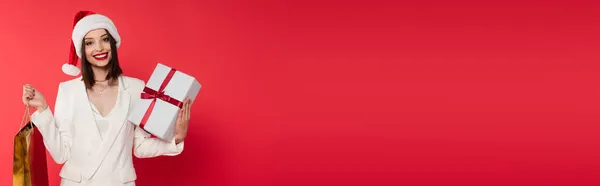 Jeune femme avec des lèvres rouges et santa chapeau tenant sac à provisions et cadeau isolé sur rouge, bannière — Photo de stock