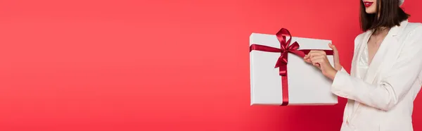 Cropped view of young woman in white jacket holding gift with bow isolated on red, banner — Stock Photo