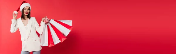 Positive woman in santa hat holding shopping bags on red background, banner — Stock Photo