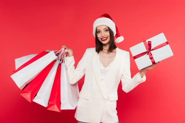 Hübsche Frau mit Weihnachtsmann-Hut, Einkaufstaschen und Geschenk auf Rot — Stockfoto