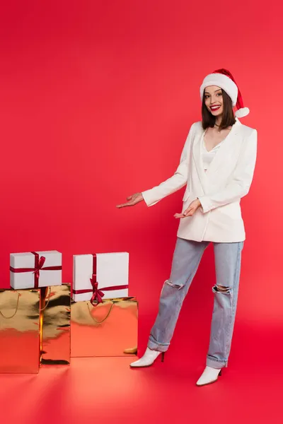 Mujer sonriente en sombrero de santa señalando regalos en bolsas de compras sobre fondo rojo - foto de stock